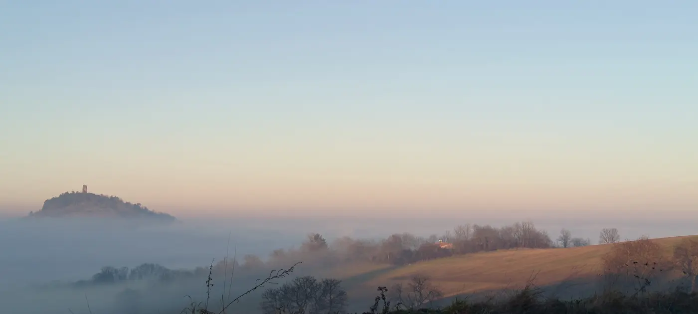 La collina con la torre, a sinistra, posata sul mare di nebbia; a destra un versante di prati già toccato dal sole radente, con le sue siepi brinate e le sue linee elettriche.