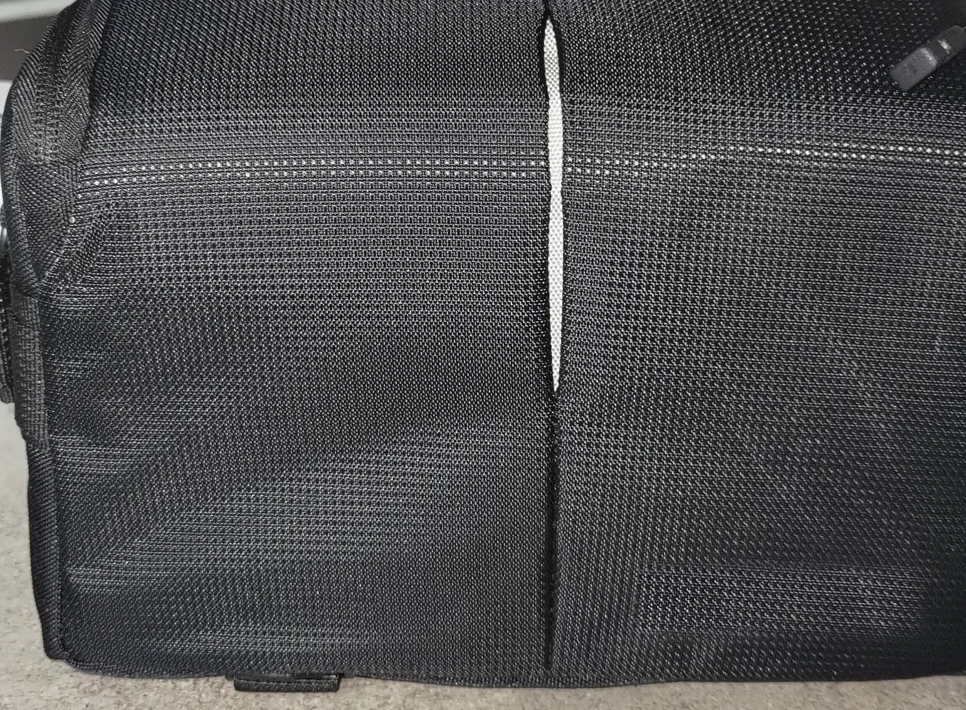 Una borsa da trasporto nera, chiusa, posata su un suolo chiaro: tela tessuta, una cucitura verticale al centro e una fettuccia riflettente lungo lo spigolo.