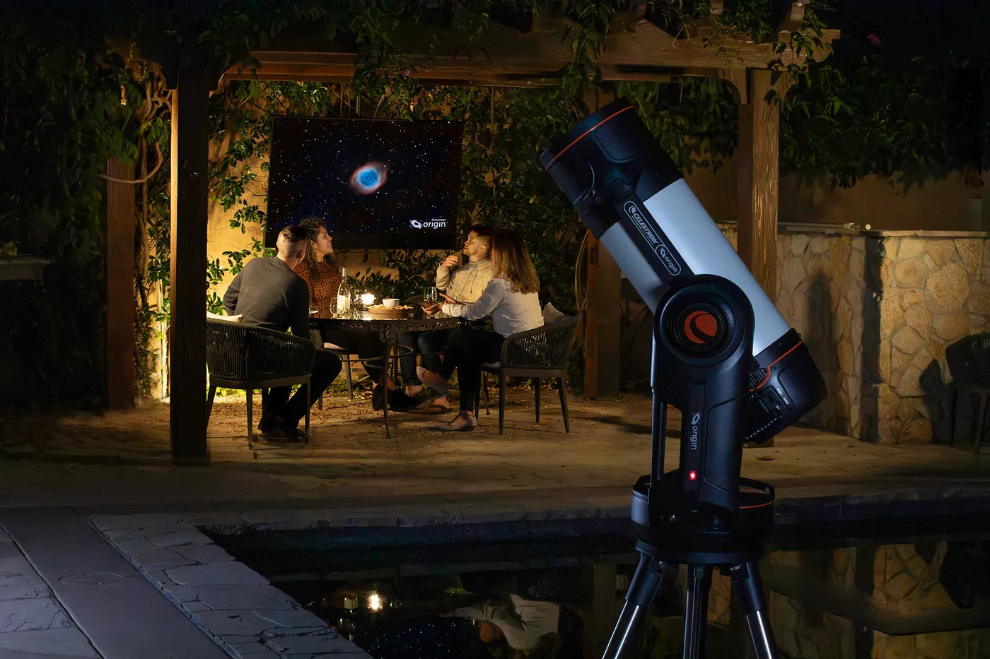 The Origin standing at the edge of a pool on a terrace, at night. Two people are sitting at a table in front of a screen in the background.