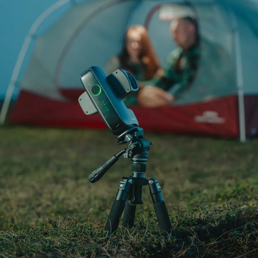El DWARF mini sobre un trípode fotográfico inclinado, clavado en la hierba delante de una tienda de campaña iluminada.