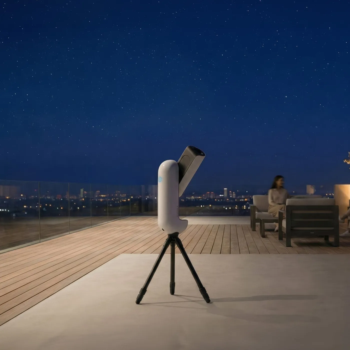 Le Vespera 3 sur son trépied au bord d'une terrasse en bois, la nuit. Les lumières d'une ville s'étirent à l'horizon et une personne est assise dans un fauteuil au fond.