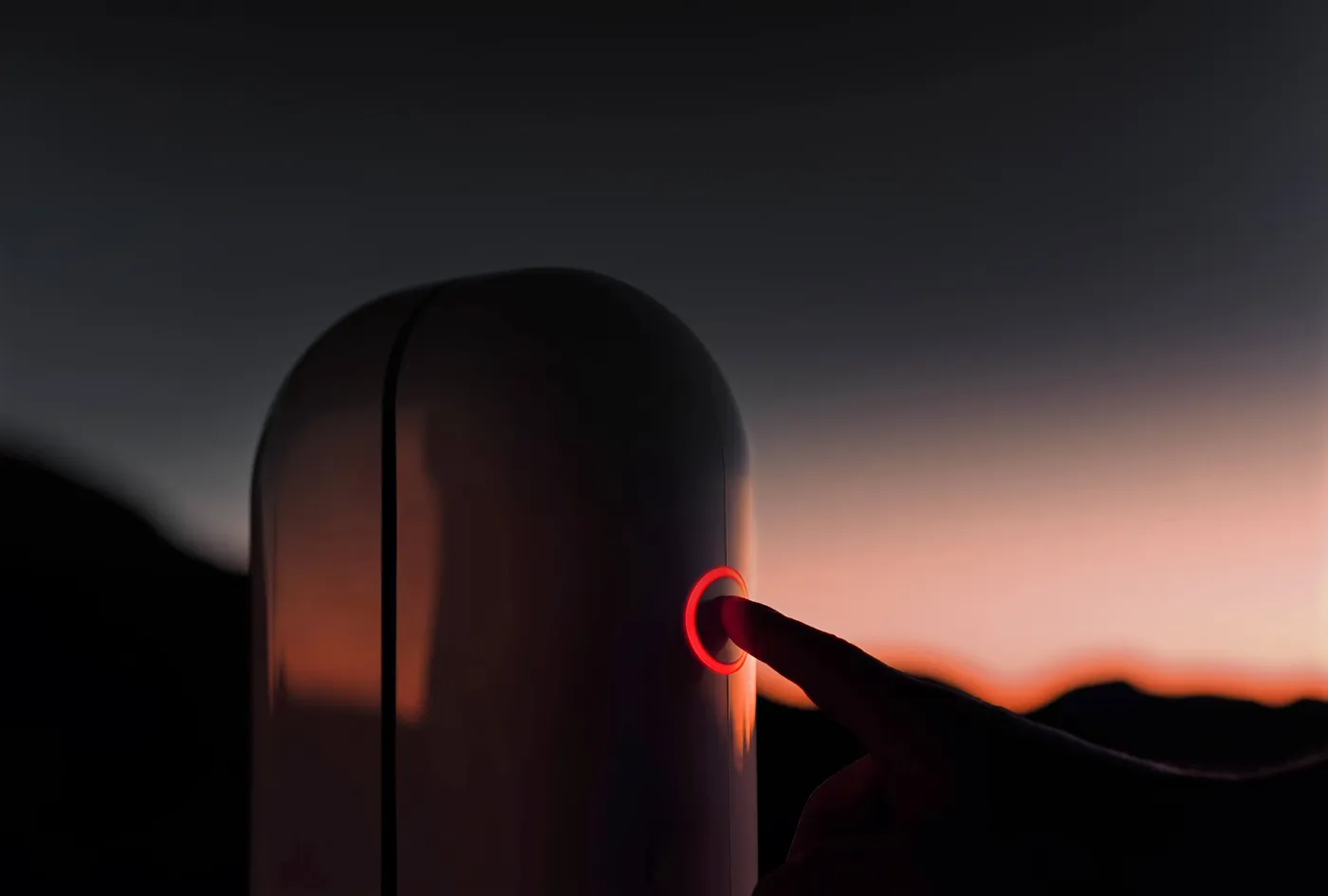 A finger presses the red lit ring of the Vespera Pro, outside at twilight, in front of an orange horizon.